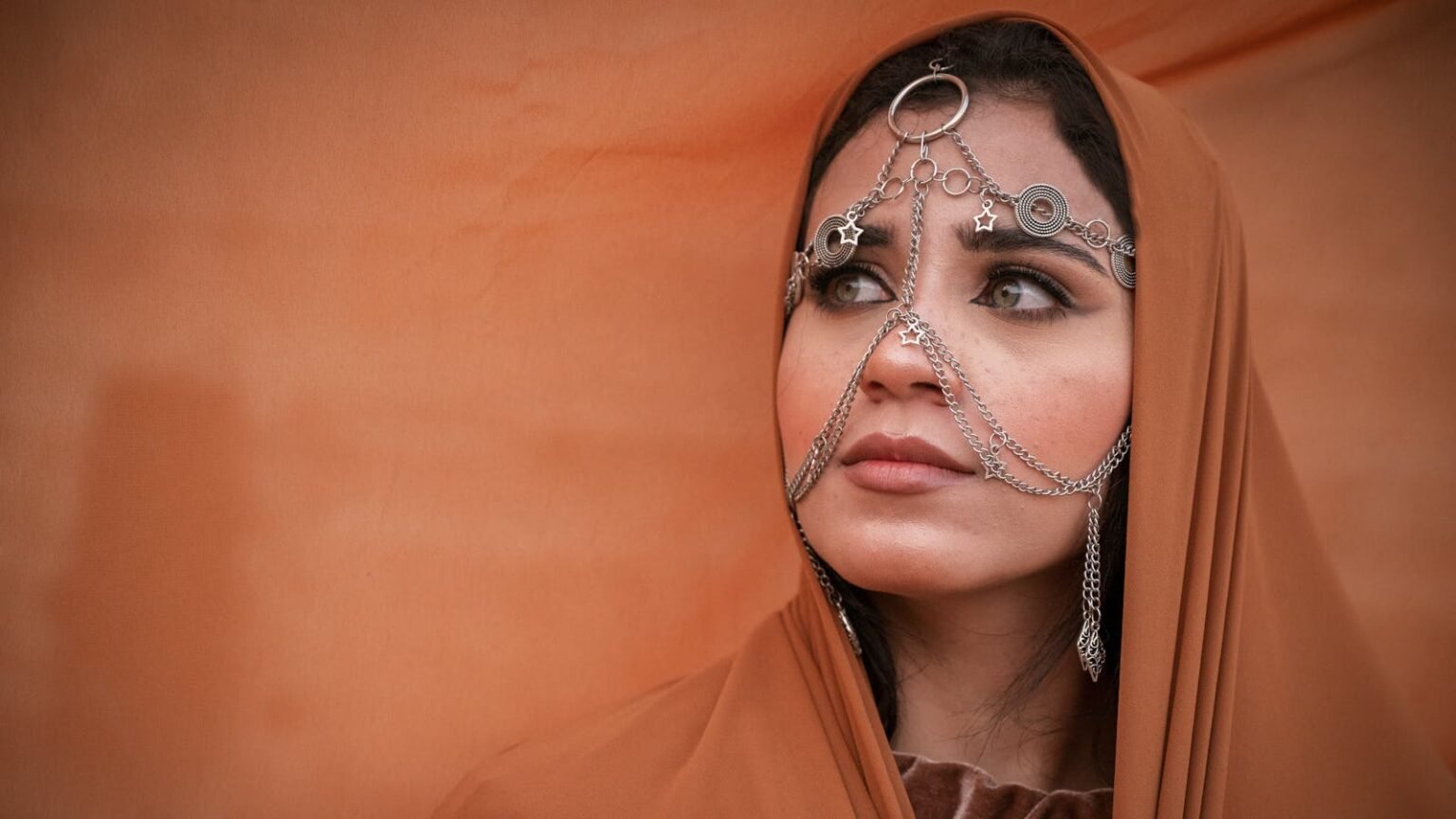 A woman in traditional attire adorned with intricate jewelry, set against a warm orange background.