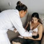 A healthcare professional helps a new mother care for her baby on a sofa during a home visit.