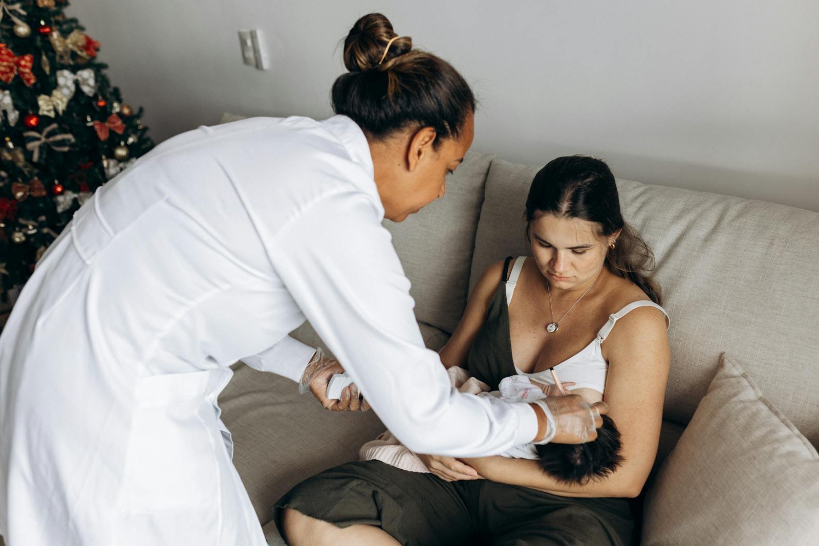 A healthcare professional helps a new mother care for her baby on a sofa during a home visit.