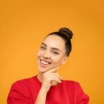 A young woman with a stylish bun poses confidently indoors with a vibrant yellow background.