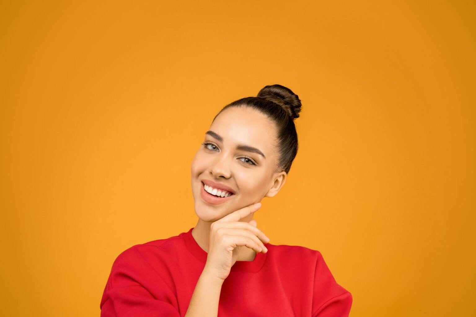 A young woman with a stylish bun poses confidently indoors with a vibrant yellow background.