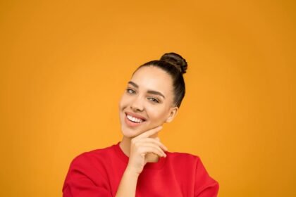 A young woman with a stylish bun poses confidently indoors with a vibrant yellow background.