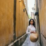 Elegant Asian woman poses in a narrow alley wearing an ao dai and holding a non la.