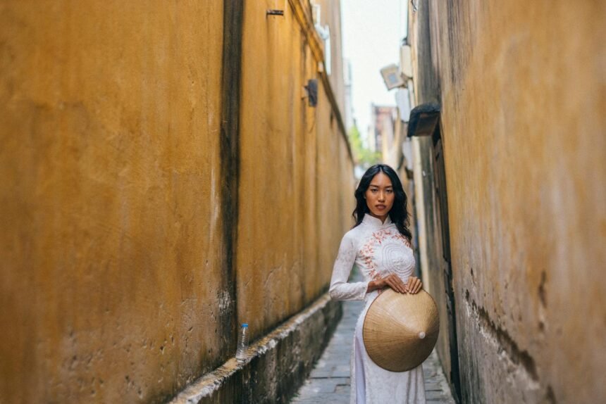 Elegant Asian woman poses in a narrow alley wearing an ao dai and holding a non la.